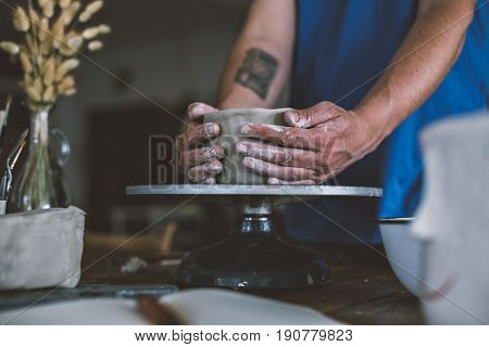 Close up of male hands working with clay on artisan working stand from dry wood surrounded by artistic workshop tools and implements creates poterry diy