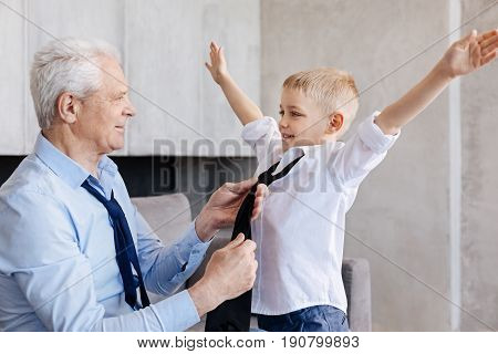 Stylish thing. Stylish pleasant delighted man teaching his grandson on how wearing good accessories while getting him ready for a school day
