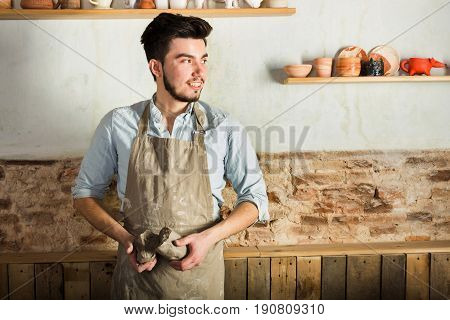 potter, tools, ceramics art concept - smiling brunette man with a beard dressed in an apron, hands of young master holding the large pieces of fireclay, a ceramist with raw materials at workshop