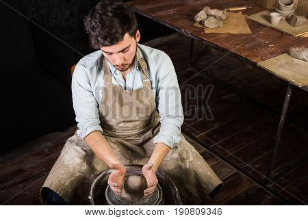 potter, workshop, ceramics art concept - young man working on potter's wheel with raw fireclay with hands, a male sculpts a utensil near wooden table with tools, master in apron and shirt, top view