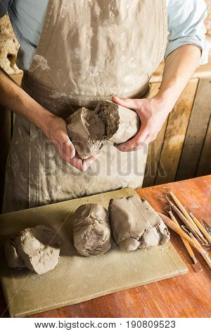 potter, clay, ceramics art concept - top view on hands of young master holding the large pieces of fireclay, male dressed in an apron, a ceramist with raw materials on table with sculpting tools set.