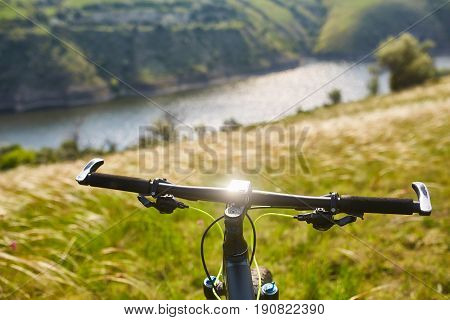 Close-up of helm of the mountain bicycle in the green grass against beautiful landscape. Detail of the bicycle and nature. Horizontal photo. Extreme travel in the countryside. Sporty backgrounds. Concept of the healthy lifestyle.