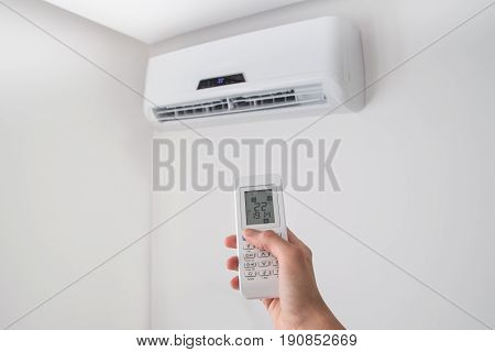 Hand holding remote control, adjusting temperature of air conditioner mounted on a white wall. Indooor comfort temperature