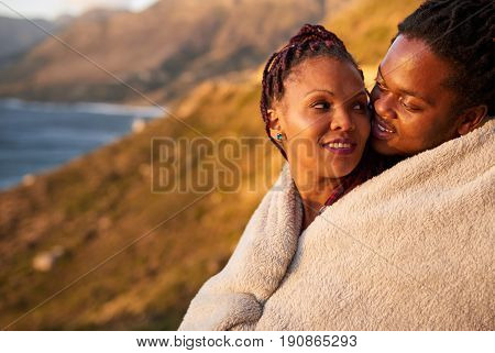 Couple keeping warm under a blanket outdoors, with a stunning view of mountains and the ocean behind them, they enjoy an inimate moment with each other by showing their affection for one and other.