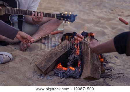 Closeup view of young and cheerful friends sitting on beach and fry sasuages or weenies in bonfire One man is playing guitar. Music on Wild beach