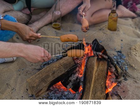Closeup view of young and cheerful friends sitting on beach and fry sasuages or weenies in bonfire One man is playing guitar. Music on Wild beach