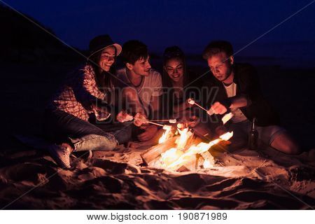 young and cheerful friends sitting on the beach and fry marshmallows near bonfire They look happy and smiling. Night time