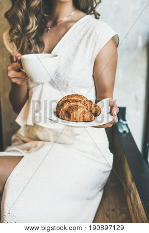 French or Italian breakfast. Young blond woman in white dress holding fresh croissant in plate and cup of cappuccino in cafe