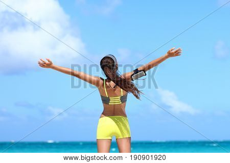 Fitness healthy lifestyle runner exercise success. Happy woman running with open arms in freedom listening to music with earphones and smartphone armband on blue copy space sky.