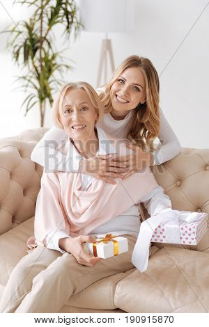 Presents for the dearest. Elegant senior lady sitting on the sofa and holding gift boxes while her beautiful young daughter standing behind the sofa and embracing the mother from