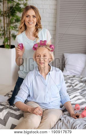 We love primping. Elegant senior woman sitting on the bed and wearing three pink hair rollers while her beautiful young daughter standing on her knees behind the mother and holding a roller in a hand