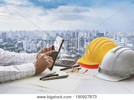 hand of business man touching on computer tablet screen and safety helmet on working table against cities of high building background