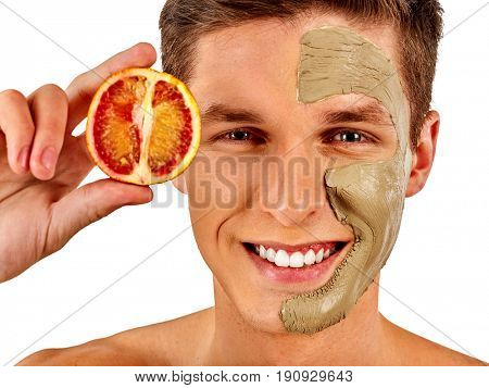 Facial mask from fresh fruits and clay for man concept. Face with treatment mud applied. Male holding lemon half for skin care procedure in salon. Man smiles dazzlingly on isolated.