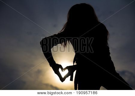 Silhouette of a young hipster woman body shape posture with hands form symbol in heart shape on sunset sky and cloud on the mountain. 