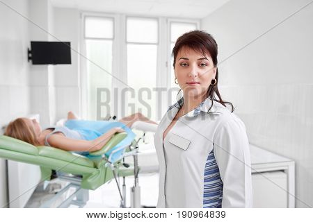 Confident female doctor posing at her clinic with female patient lying on gynecological chair on the background waiting for medical examination. Health medicine concept.