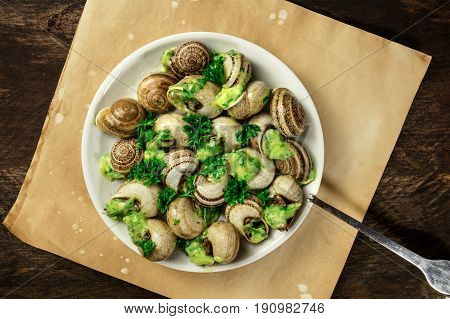 An overhead photo of escargots a la bourguignonne, a plate of snails with the typical French green sauce of garlic and herb butter, on a rustic texture with a place for text and a special fork