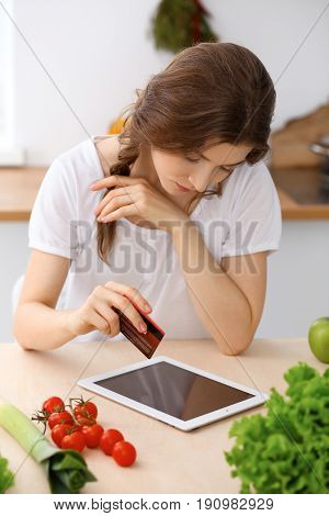 Young woman looking for a new recipe for cooking in a kitchen. Housewife is making online shopping by tablet computer and credit card.