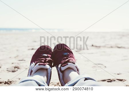 Feet Of Young Woman In Sport Footwear And Jeans Relaxing After Hiking On Sand Beach. Pov. Adventure 