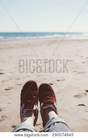 Feet Of Young Woman In Sport Footwear And Jeans Relaxing After Hiking On Sand Beach. Pov. Adventure 