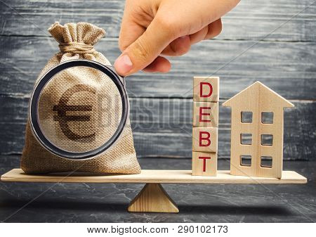 Money Bag And Wooden Blocks With The Word Debt And A Miniature House On The Scales. Payment Of Debt 