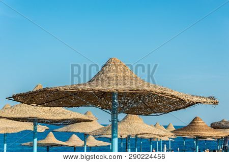 Lonely Wicker Sun Umbrella At Mediterranean Beach By Sea. Natural Bamboo Sunshades And Summer Umbrel