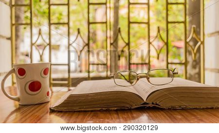 An Open Book A Coffee Cup And A Specs Or Eyeglass On Wooden Table, Warm Sunny Morning, Side View Clo