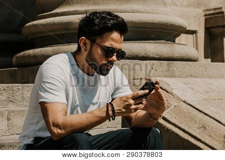 Handsome Bearded Hispanic Man Using Mobile Smartphone For Surfing Web Internet, Making Call And Look