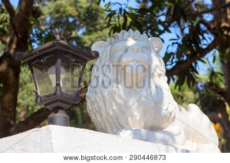 A White Sculpture Of A Lying Lion In The Park Guards A Latern.