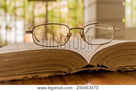 An Open Book And A Eyeglass Nicely Placed On Wooden Table On A Warm Sunny Morning, Side View Close U