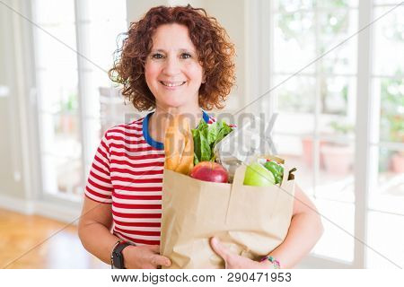 Senior woman holding paper bag full of fresh groceries from the supermarket with a happy face standing and smiling with a confident smile showing teeth