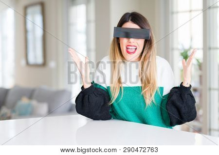 Young beautiful woman blindfold eyes with black censor band at home celebrating mad and crazy for success with arms raised and closed eyes screaming excited. Winner concept