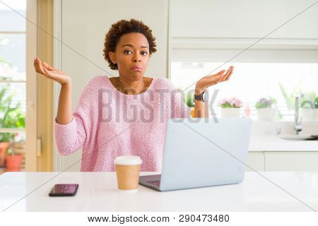 Young african american woman working using computer laptop clueless and confused expression with arms and hands raised. Doubt concept.