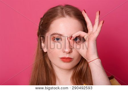 Studio Shot Of Attractive Serious Young Woman, Makes Ok Sign, Covers Her Eye, Expresses Confidence, 