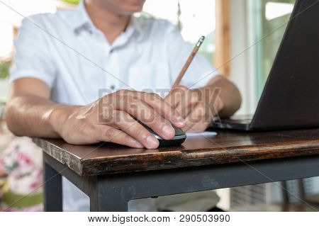 Asian Tan Skin Holding Wireless Mouse On The Vintage Wood Table And Writting Pencil With His Left Ha