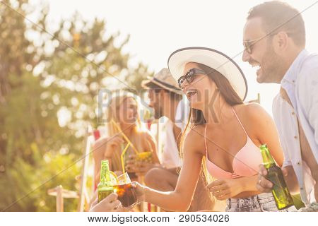 Group Of Friends At A Poolside Summer Party, Sitting At The Edge Of A Swimming Pool, Drinking Cockta
