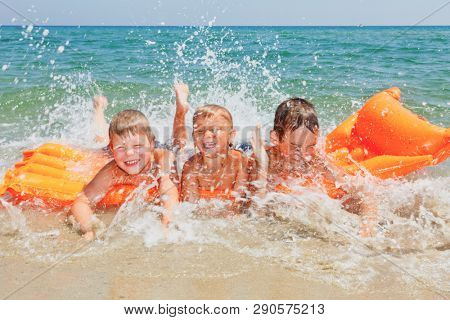 Happy children having fun on a beach splashing water laying on inflatable mattress enjoying summer vacation, clear sea water is seen in background