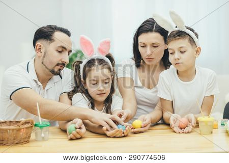 Inspired Family In White T-shirts Sitting Together At The Table, Holding Colored Easter Eggs In Thei