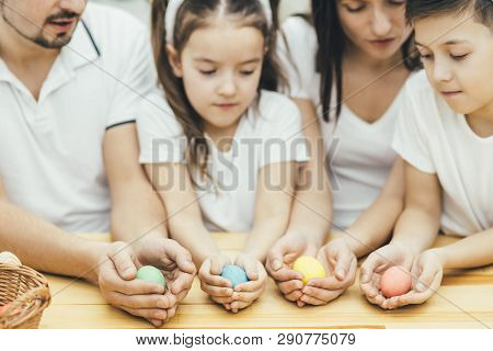 Inspired Family In White T-shirts Sitting Together At The Table, Holding Colored Easter Eggs In Thei