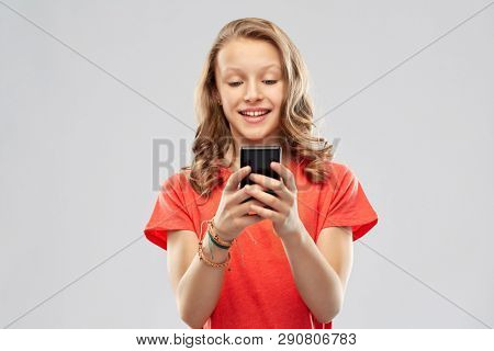 technology and people concept - smiling teenage girl in red t-shirt using smartphone over grey background
