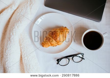 Big White Morning Cup Of Coffee And Crossant In Bed With Notebook, Glasses, Pillow And Cosy Blanket.