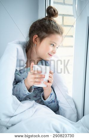A little girl sittiing on a windowsill on a pillow covered with a blanket. Childhood. Kid's fashion.