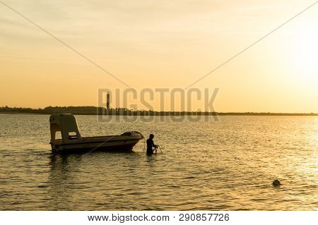 Silhouette Of A Man Taking Anchor For A Boat With Setting Sun In The Distance