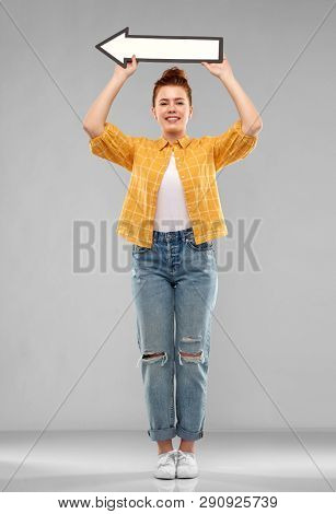 direction and people concept - smiling red haired teenage girl in checkered shirt holding big white leftwards thick arrow over grey background