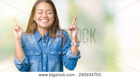 Young beautiful brunette woman wearing blue denim shirt over isolated background smiling crossing fingers with hope and eyes closed. Luck and superstitious concept.