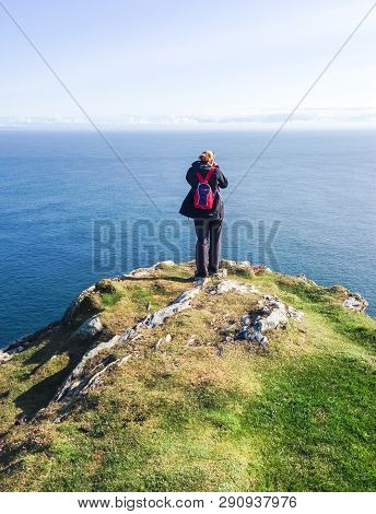 A Hiker With A Small Red Backpack Stands On The Edge Of A Cliff Overlooking The Ocean On A Sunny Day