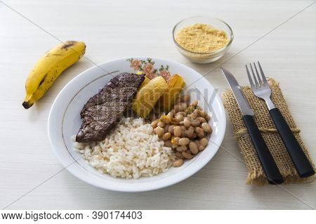 Plate Of Rice, Beans, Steak And Cassava. With Cutlery And Manioc Flour And Banana For Dessert. White