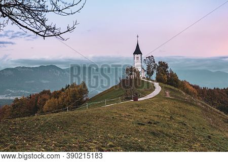 Beautiful Sunset Landscape Of Church Jamnik In Slovenia On Green Hill With The Trees And Pink Sky