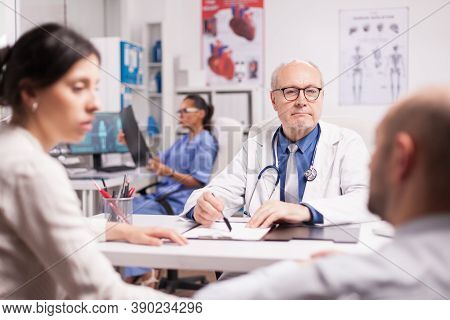 Senior Doctor During Consultation With Disabled Young Man In Wheelchair Accompanied By Wife. Medic W