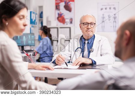 Older Doctor Looking At Disabled Man In Wheelchair During Consultation In Hospital Cabinet. Handicap
