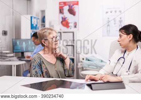 Senior Woman Discussing About Neck Pain With Doctor During Consultation In Hospital Office. Medic We